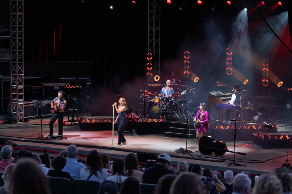 Grammy-nominated band Lake Street Dive performs onstage under vibrant red geometric lighting as a seated crowd watches. The lead singer stands at the microphone, flanked by band members playing guitar, drums, and keyboards.