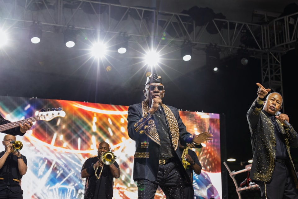 Earth, Wind & Fire performs on stage in gold-sequined suits. The lead singer is centered, with a vocalist to his right pointing to the crowd, while a brass section plays behind them.