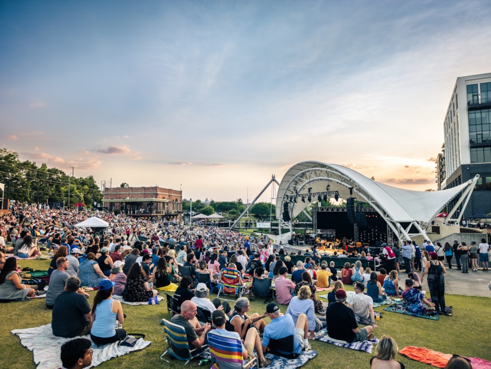 A large crowd relaxes on the grassy hillside with blankets and lawn chairs while Waxahachee performs onstage under the canopy of the Adderley Amphitheater.